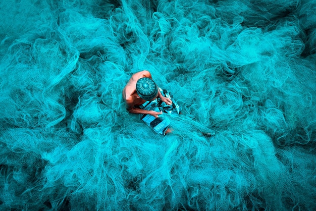 Old fisherman hands sewing blue fishing nets sitting on the ground and surrounded big net