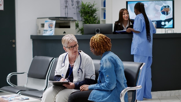 Old general practitioner taking notes at consultation with patient, doing medical checkup visit with african american person in hospital waiting room. Doctor and woman talking about diagnosis.