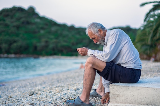 An old man on the beach in a white shirt and dark pants An aged man with a beard walks alone on the beach at sunset Portrait of a lonely grayhaired old man Palm trees on the background