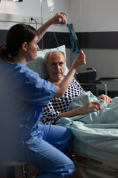 Old patient with lungs illness breathing using oxygen mask laying in hospital bed, listening nurse showing x-ray explayning diagnosis before surgery