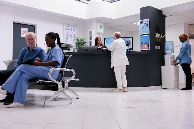 Old physician working with asian receptionist to plan checkup report papers at hospital reception desk. Medic taking to clinic worker at counter about medical forms and healthcare insurance support.