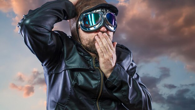 Old plane pilot on background of storm clouds with expressive face. glasses and old hat with black leather jacket