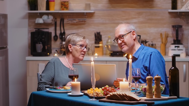 Old retired senior couple using laptop in the kitchen. Elderly people sitting at the table browsing, searching, using laptop, technology, internet, celebrating their anniversary in the dining room.