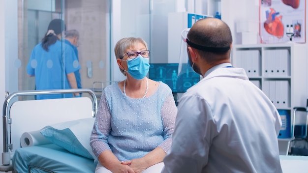 Old retired senior woman wearing a mask at doctor consultation during COVID-19 pandemic. Healthcare system, disease treatment in modern professional private clinic or hospital