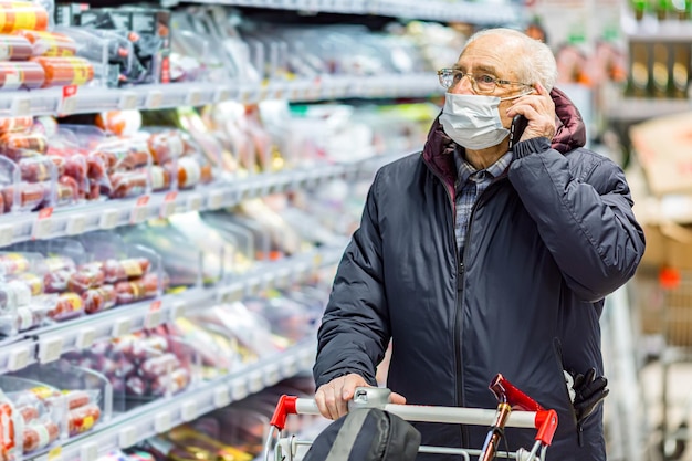 Old senior european man wearing protective facial mask pushing shopping cart and talking on the phone in the supermarket. Shopping during COVID-19 concept.