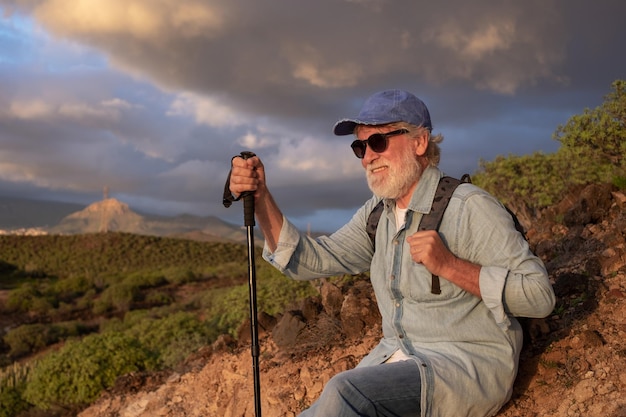 Old senior man in hat and casual clothing sitting in outdoor excursion at sunset light enjoying freedom and vacation Caucasian bearded male with stick looking the mountain tropical landscape