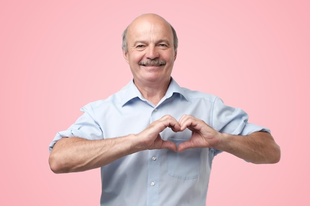 Old senior man with mustache making out of hands heart over pink background