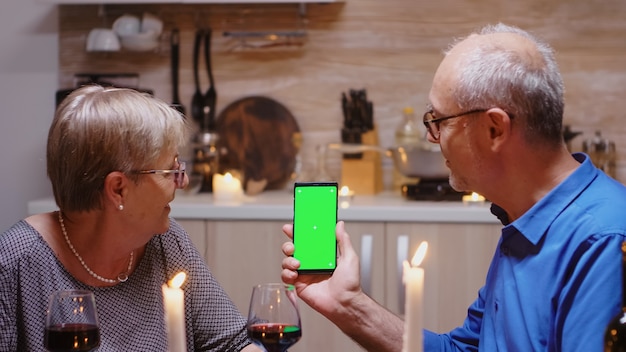 Old senior retired couple holding green screen phone at dinner. Aged people looking at mockup template chroma key isolated smart phone display using techology internet sitting at the table in kitchen.