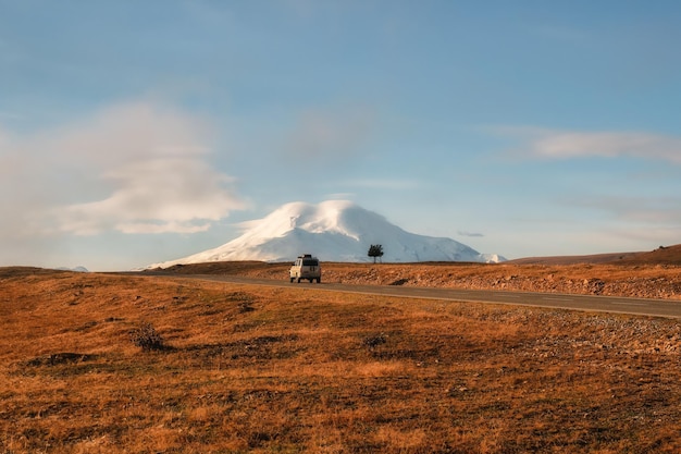 Old silver SUV is driving along the highway route to the dawn snowcovered Elbrus Autumn beautiful road through the mountain desert Morning mountain valley with a serpentine road