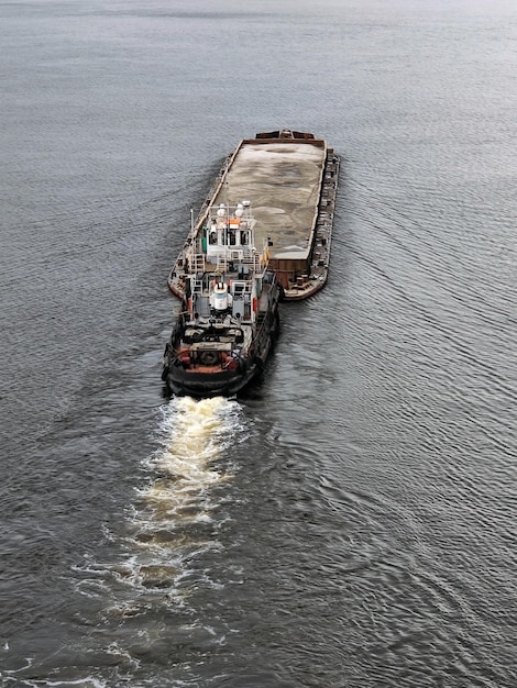 An old tugboat pushes a barge of sand in front of it and floats down the river View from above