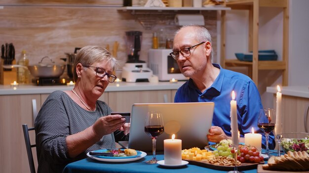 Old woman paying online the holiday using laptop and card during romantic dinner. Old people sitting at the table, browsing, using the technology, internet, celebrating their anniversary.