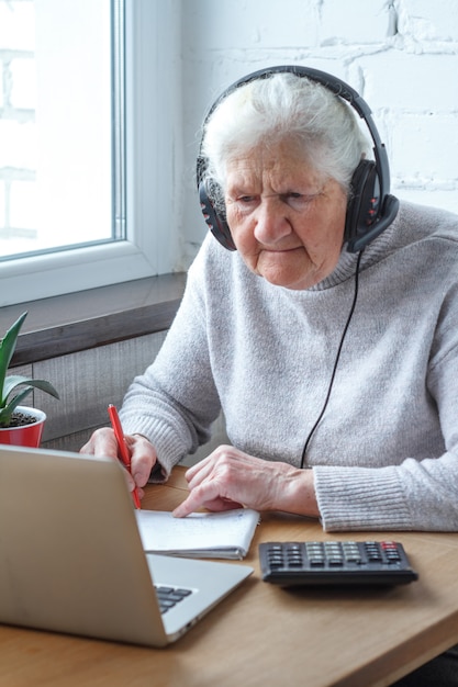 An old woman sits at a table in front of a laptop with headphones and writes into a notebook.