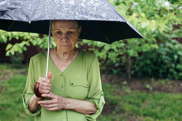 An old woman stands on the street under the rain with a black umbrella