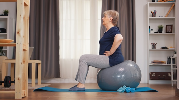 Old woman workout on training ball in living room. Old person training home healthy retirement lifestyle stretching, home training and exercise