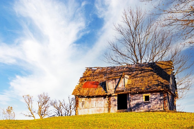 Old wooden house built on grassy hill top near bare trees in highland against mountains under blue sky with white cirrus clouds in autumn