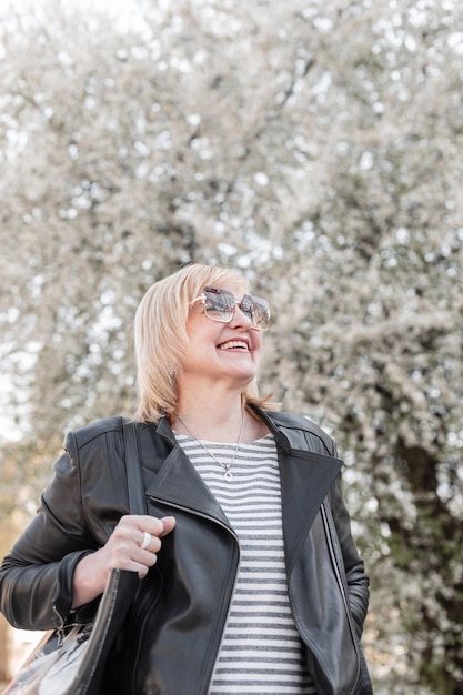 An older beautiful fashion happy woman with a smile and trendy sunglasses in a fashionable black leather jacket with a polo shirt and handbag enjoy and walking near a flowering tree on the street