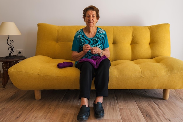 Older caucasian woman knitting looking at camera smiling Sitting on a yellow sofa