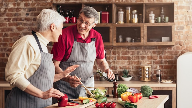 An older couple in a modern kitchen wearing aprons prepare a fresh salad together the man is pouring