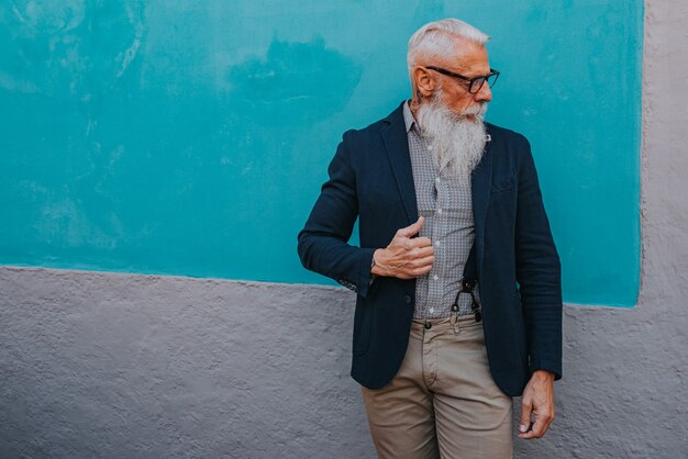 An older man in hipster clothes and glasses and a long white beard poses on a blue wall