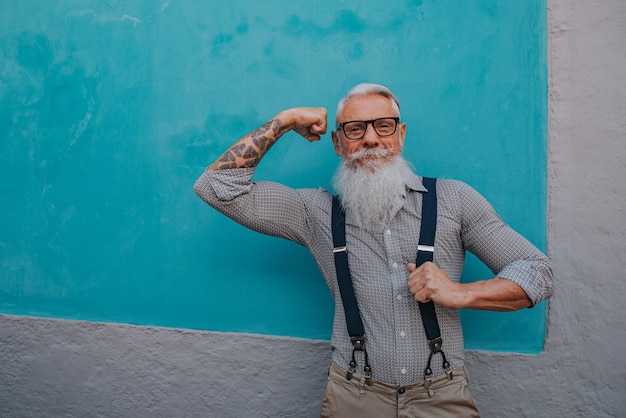 An older man in hipster clothes and glasses and a long white beard poses on a blue wall