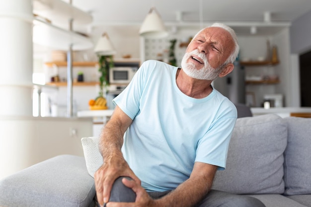 The older man is sitting on the couch at home has pain in the knee joint holding his leg osteoarthritis concept