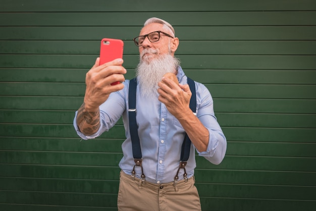 An older man older man in hipster clothes and glasses and a long white beard comb his beard and take a photo