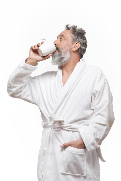 Older man with beard in bathrobe drinking a cup of coffee in with white background