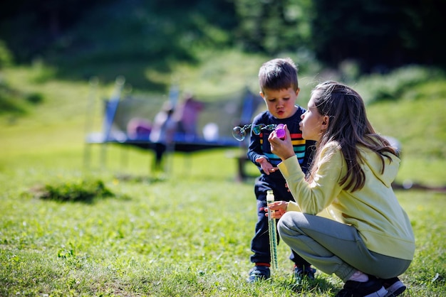 Older sister plays with younger brother and teaches him how to blow soap bubbles on the backyard in