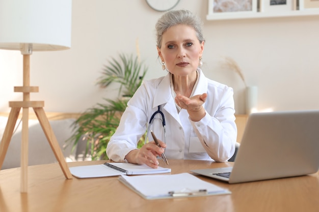 Older woman doctor therapist wearing headset video call talking to web camera consulting virtual patient online by video conference call chat.