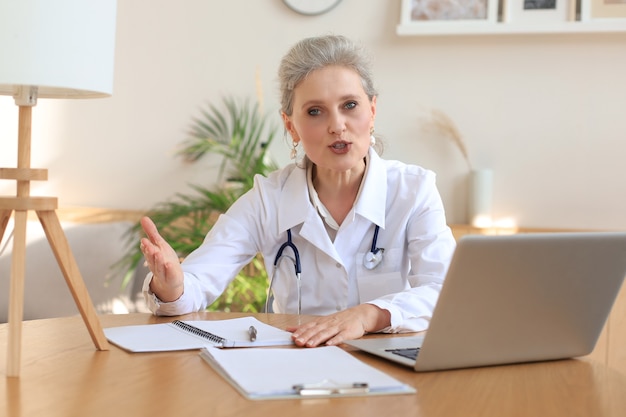 Older woman doctor therapist wearing headset video call talking to web camera consulting virtual patient online by video conference call chat.