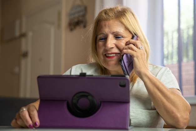 Older woman sitting at home while talking on her cell phone with someone else and is entertaining herself with her tablet