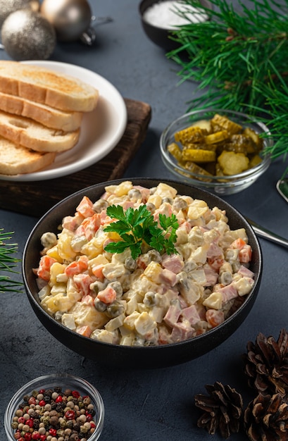 Olivier salad in a black bowl on a dark background with Christmas decorations. Side view, vertical.