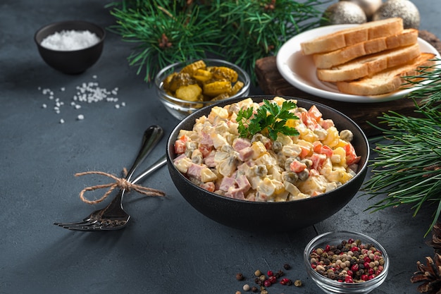 Olivier salad in a black bowl on a dark background with pine branches. Side view, copy space.