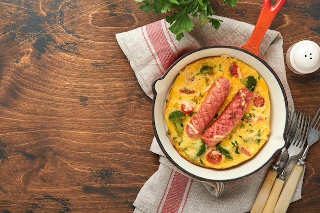 Omelette or frittata with two sausages melted cheese cherry tomatoes broccoli and parsley on red skillet or iron pan on old wooden rustic table background Healthy breakfast food concept Top view