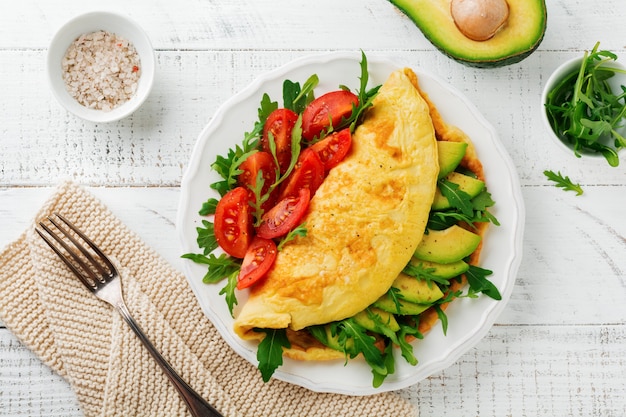 Omelette with avocado, tomatoes and arugula on white ceramic plate on light stone background