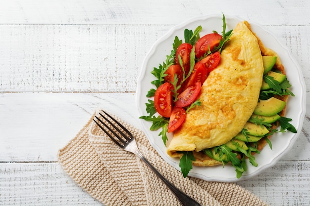 Omelette with avocado, tomatoes and arugula on white ceramic plate on light stone surface. Healthy breakfast. Selective focus. Top view.