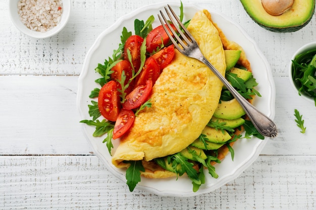 Omelette with avocado, tomatoes and arugula on white ceramic plate on light stone surface