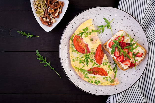 Omelette with tomatoes, ham, green onion and sandwich with strawberry on dark table