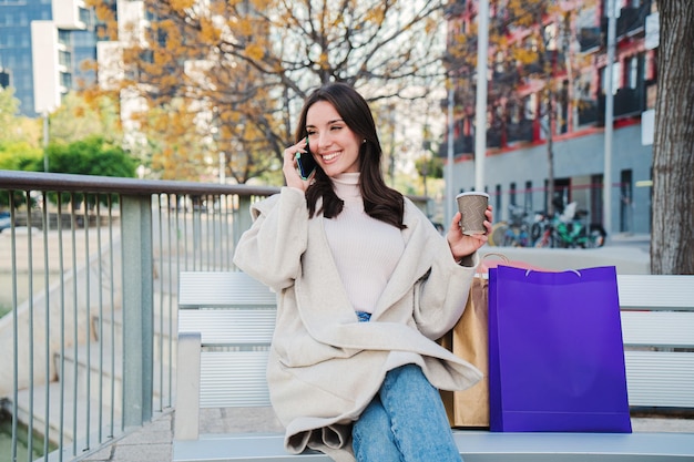 One brunette lady doing a call with a cell phone sitting on a bench in a city street after shopping