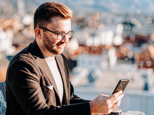 One caucasian man wearing glasses holds smartphone and drinks coffee from cup at cafe. Remote work via smart phone. City life. Business person at break. Casual fashion. Success concept. Mixed media.