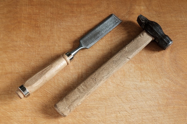 One chisel with a wooden handle and an old hammer with a wooden handle lie diagonally on a wooden table.