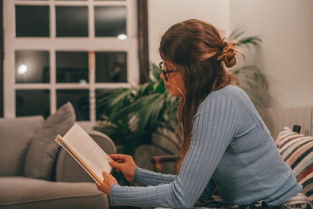 One cute and attractive wearing eye glasses reading and looking an interesting book at home indoor on the sofa Young woman relaxing alone at night reading books sitting on the couchxA