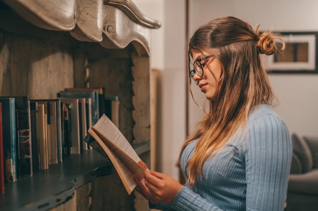 One cute and attractive wearing eye glasses reading and looking an interesting book at home indoor Young woman relaxing alone at night reading books