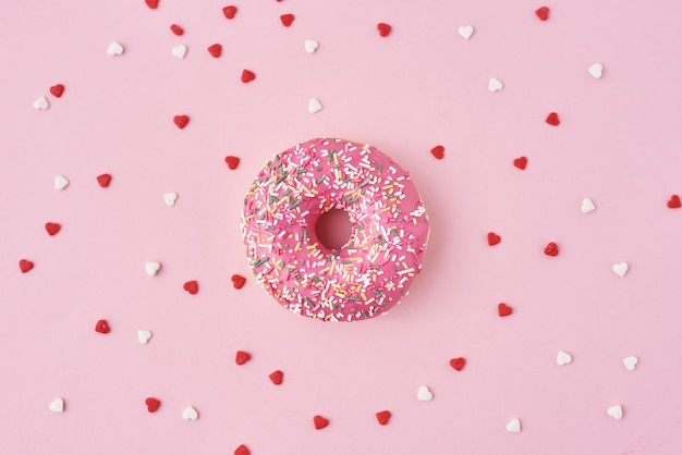 One donuts decorated icing and sprinkles and confetti in shape of heart on pink background