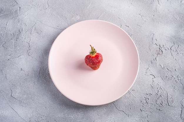 One fresh ripe strawberry fruit in pink plate, summer vitamin berries on grey stone table, top view