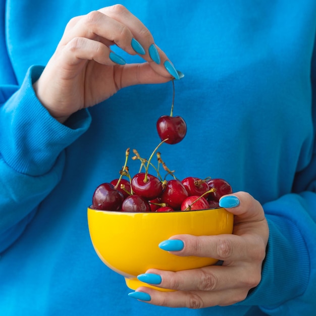 One hand holds a yellow bowl with cherries the other holds one berry by the stem