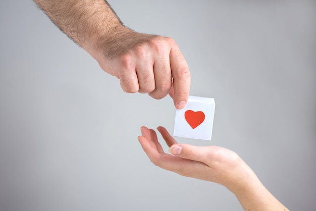 One male hand gives to one female hand a white box with a heart, simbol of love, close up.