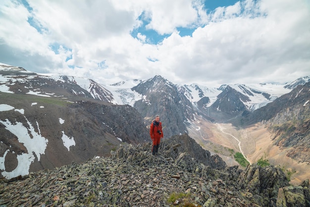 One man in red with camera near precipice edge with sharp rocks with view to large snowy mountains and glacier under cloudy sky Tourist on stone hill with view to mountain range at changeable weather