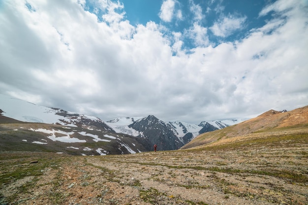 One man walks with view to high snowy mountain range with sharp tops and glaciers under gray cloudy sky Colorful landscape with tourist on background of large snow mountains at changeable weather