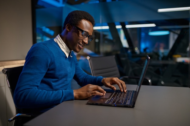 One manager in glasses works on laptop in night office. Male worker, dark business center interior, modern workplace